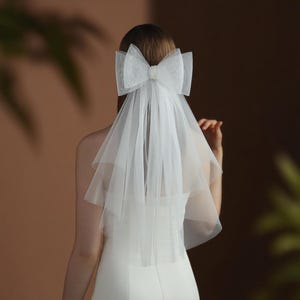 Könnte beinhalten: Ein weißer Brautschleier mit einer großen Schleife und einem Perlenakzent. Der Schleier besteht aus einem transparenten, mehrschichtigen Stoff, der über den Rücken fällt. Die Schleife ist der Blickfang und verleiht dem Brautensemble einen Hauch von Eleganz.