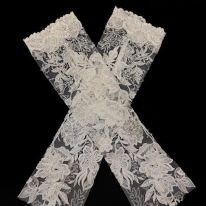 May include: Two white lace arm cuffs with floral and leaf patterns. The cuffs are embellished with sequins and beads, and are set against a black background. The lace has a delicate, intricate design.