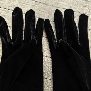 May include: A pair of black velvet gloves featuring a soft, light-reflecting texture. The gloves are designed with individual finger compartments and extend past the wrist, showcasing a deep, solid black color throughout the entire fabric surface.