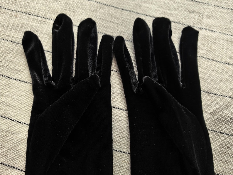 May include: A pair of black velvet gloves featuring a soft, light-reflecting texture. The gloves are designed with individual finger compartments and extend past the wrist, showcasing a deep, solid black color throughout the entire fabric surface.