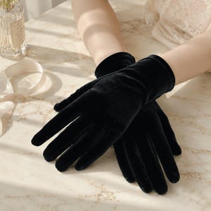 May include: A pair of solid black velvet gloves with a smooth, soft-touch finish. The gloves feature a classic wrist-length cut, providing full coverage for the fingers and hands with a consistent, deep black color throughout the fabric.