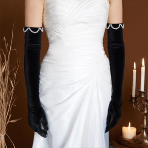 Peut inclure: Longues gants d'opéra en velours noir, ornés d'une bordure de perles sur le haut du bras. Les gants s'étendent au-delà du coude. L'arrière-plan présente une robe blanche et des bougies allumées, pour un événement formel ou un mariage.