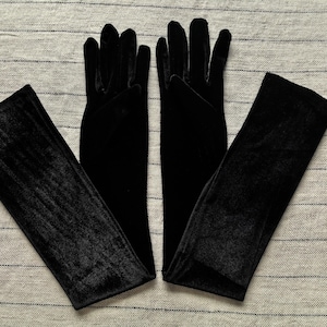 May include: A pair of long, black velvet gloves that extend past the wrist. The gloves feature a soft, matte texture and are designed to cover the hands and forearms completely, with individual finger slots and a smooth, seamless appearance.