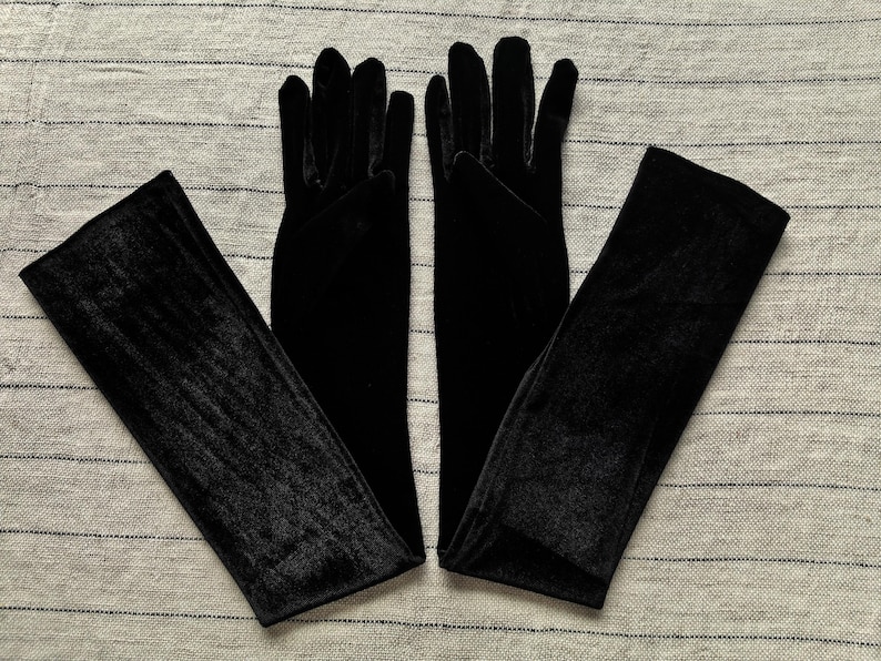 May include: A pair of long, black velvet gloves that extend past the wrist. The gloves feature a soft, matte texture and are designed to cover the hands and forearms completely, with individual finger slots and a smooth, seamless appearance.
