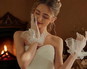May include: Delicate white sheer gloves with ruffled cuffs and pearl embellishments. The gloves are made of a soft, flowing fabric. These gloves are a bridal accessory, perfect for a wedding.