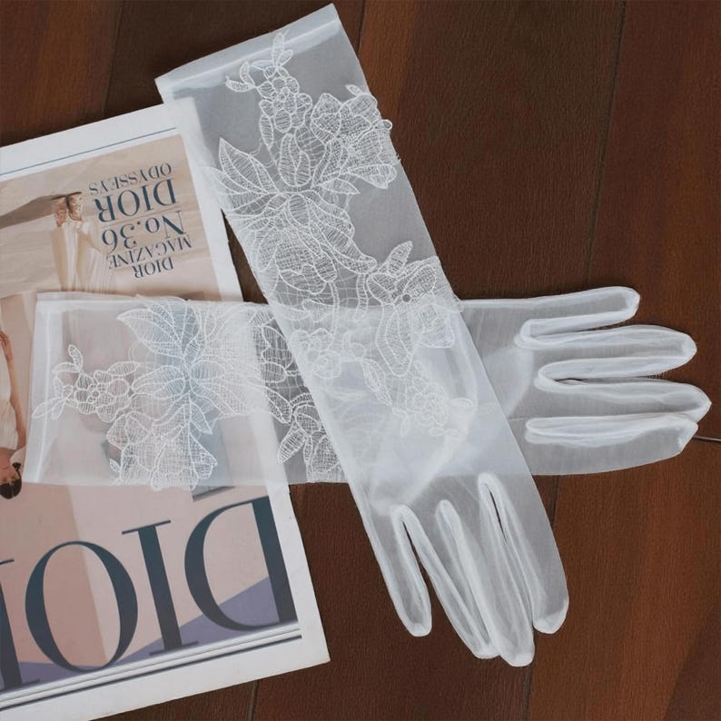 May include: A pair of sheer white gloves with intricate floral lace detailing. The gloves are long, extending past the wrist, and are made of a delicate, translucent fabric. The gloves are laid out on a wooden surface next to a magazine.