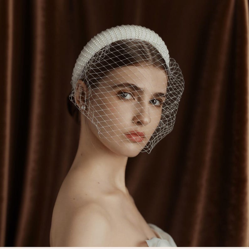 May include: A white beaded headband with a delicate net veil. The headband is thick and covered in small, evenly spaced beads. The veil is a fine, diamond-patterned mesh, draped over the face. The background is a solid, dark brown.