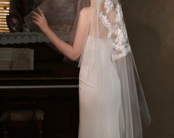 May include: A bride wearing a white strapless gown and a sheer veil with floral lace edging. The veil is draped over the head and shoulders. The background features a piano and a vintage painting, creating a romantic scene.
