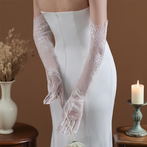 May include: Elegant white lace gloves, extending to the upper arm, with intricate floral patterns. The gloves are sheer, revealing the skin beneath, and are paired with a white dress. A classic accessory for weddings or formal events.