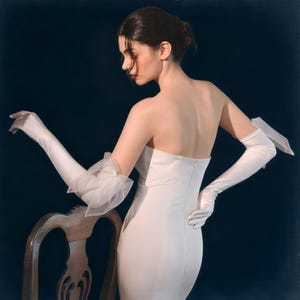 Op de afbeelding: Een strapless witte jurk met een getailleerd silhouet. Het model draagt lange, witte satijnen handschoenen, één met een doorschijnend, gerimpeld accent bij de elleboog. De achtergrond is donker.