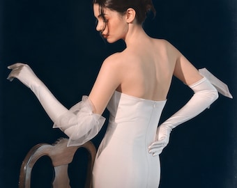 May include: A strapless white gown with a fitted silhouette. The model wears long, white satin gloves, one with a sheer, ruffled accent at the elbow. The background is a dark, solid color.