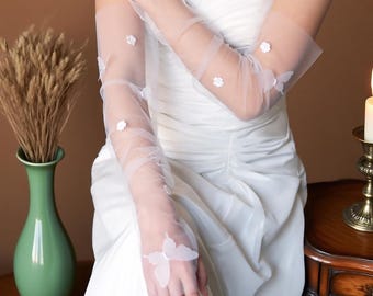 May include: Sheer white bridal gloves adorned with delicate white butterfly and flower appliqués. The gloves are long, extending past the elbows, and are made of a lightweight, translucent fabric. The gloves are worn with a white strapless wedding dress.