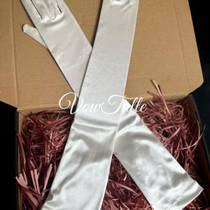 Könnte beinhalten: Ein Paar elfenbeinfarbene Satinhandschuhe, einer lang und einer kürzer, liegen in einer offenen Pappschachtel mit roten Papierstreuseln. Die Handschuhe haben eine glatte, glänzende Textur und eignen sich für formelle Anlässe.