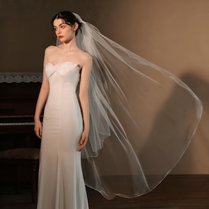May include: A strapless white wedding dress with a flowing skirt and a long, sheer veil. The veil is attached to the back of the head and cascades down the back. The dress has a fitted bodice and a simple, elegant design.