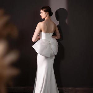 May include: A strapless white wedding gown with a fitted bodice and a large bow detail at the back. The gown has a mermaid silhouette, flowing to the floor. The model is turned to the side, with her hair up, against a dark background.