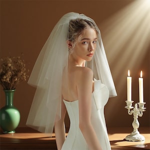 May include: A bride wearing a white strapless wedding dress and a sheer, short veil. Two lit candles in a decorative holder are to the right. A green vase with dried flowers is on the left.