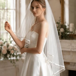 Op de afbeelding: Een bruid draagt een witte strapless trouwjurk en een doorschijnende sluier. De sluier is afgezet met fijn wit kant en kleine, bungelende kralen. De achtergrond omvat een raam en een decoratieve spiegel.