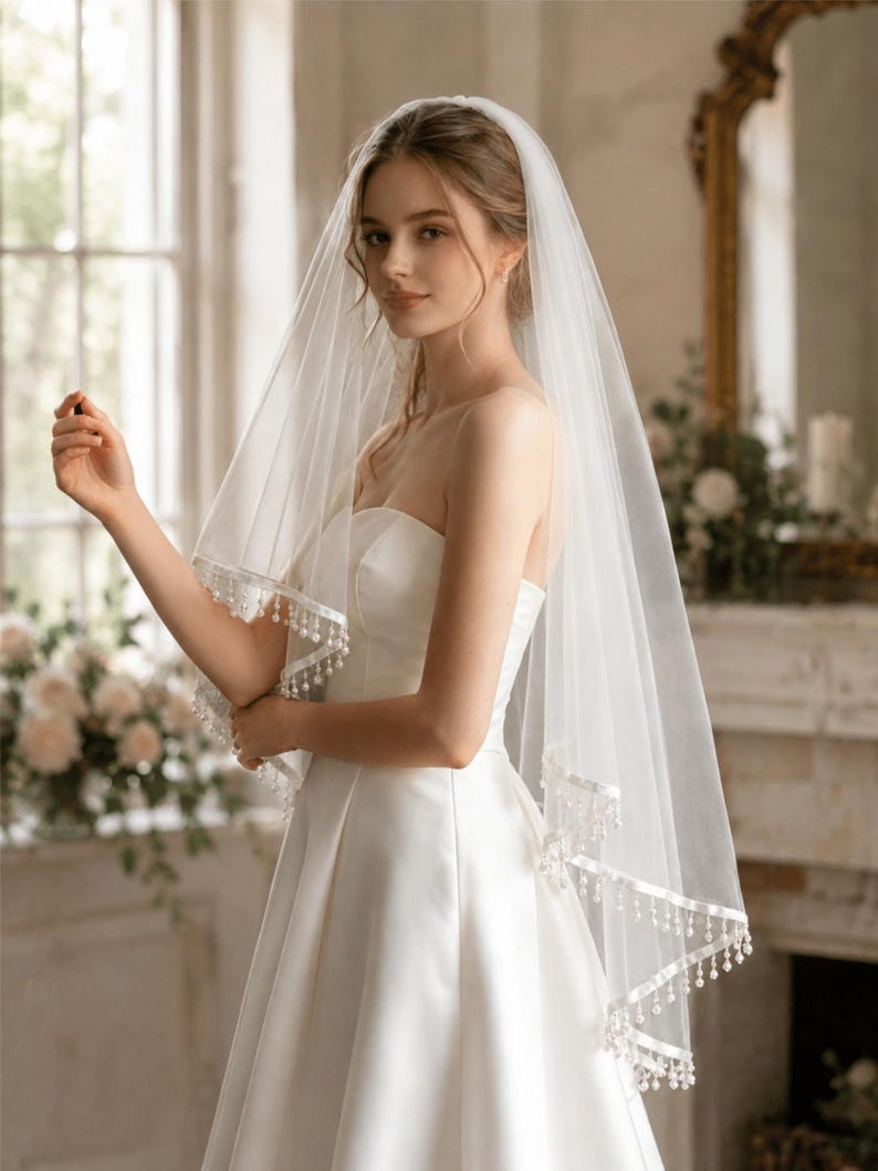 Op de afbeelding: Een bruid draagt een witte strapless trouwjurk en een doorschijnende sluier. De sluier is afgezet met fijn wit kant en kleine, bungelende kralen. De achtergrond omvat een raam en een decoratieve spiegel.