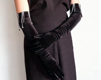 May include: Black velvet opera gloves with rhinestone fringe detail at the wrist. The long gloves are gathered along the arm and extend past the elbow. The gloves are shown against a black dress.