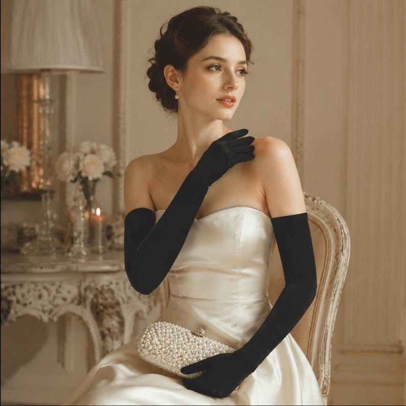 May include: A pair of long black opera-length gloves made of a smooth, matte fabric, extending past the elbows. The gloves are worn with a strapless, cream-colored satin gown and a small, rectangular clutch bag covered in white faux pearls.