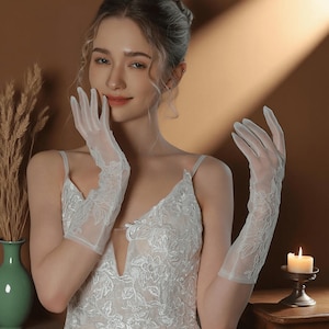 May include: Elegant white sheer gloves with floral lace detailing. The gloves are long, extending past the wrist, and are paired with a white lace dress. The image is set against a warm, neutral background, suggesting a bridal or formal occasion.