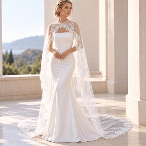 May include: A floor-length white satin gown featuring a strapless bodice and a fitted silhouette that flares at the hem. A sheer, floor-length cape with intricate white floral lace embroidery covers the shoulders and drapes down the back.