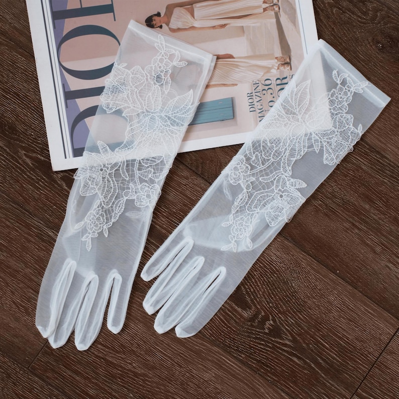 May include: A pair of sheer white gloves with intricate floral lace detailing. The gloves are long, extending past the wrist, and are laid out on a magazine with the word "DIOR" visible. The gloves are a delicate accessory.