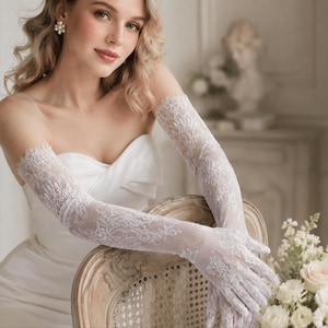 May include: A pair of long, white lace gloves featuring an intricate floral pattern that extends from the fingers up to the mid-forearm. The delicate, sheer fabric is detailed with scalloped edges and a fine mesh texture, providing a refined appearance.