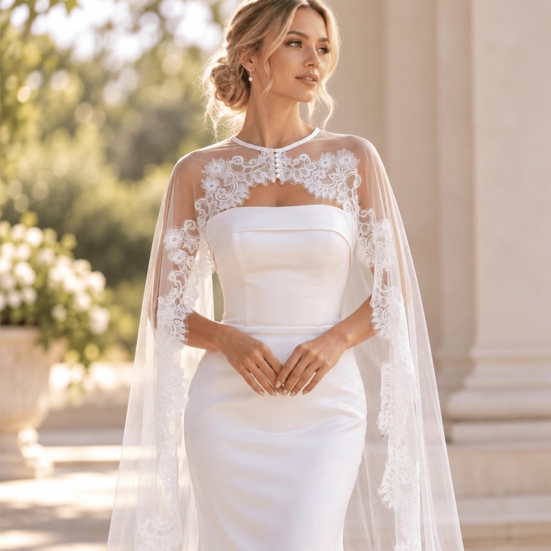 May include: A floor-length white satin gown featuring a strapless bodice and a sheer tulle cape overlay. The cape is adorned with intricate white floral lace embroidery along the neckline and edges, secured at the neck with a row of small buttons.