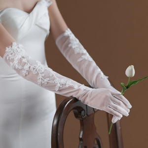 Könnte beinhalten: Elegante weiße transparente Handschuhe mit floralen Spitzendetails. Die Handschuhe reichen bis zum Oberarm und sind mit zarten gestickten Blumen und Blättern verziert. Eine weiße Tulpe wird in der Hand gehalten.