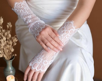 Könnte beinhalten: Weiße Brauthandschuhe aus Spitze mit zartem Blumenmuster. Die fingerlosen Handschuhe reichen bis zum mittleren Unterarm und haben einen gewellten Rand. Sie bestehen aus einem transparenten, offen gewebten Stoff.