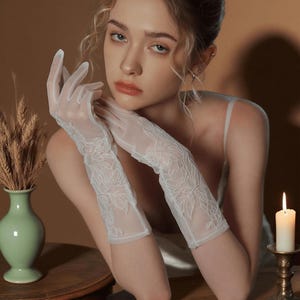 May include: Elegant white lace gloves with floral embroidery. The gloves are sheer and reach the mid-forearm. The image also features a green vase with wheat stalks, a lit candle, and a woman.