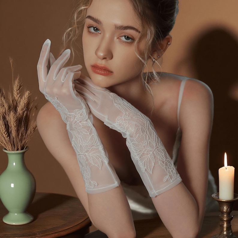 May include: Elegant white lace gloves with floral embroidery. The gloves are sheer and reach the mid-forearm. The image also features a green vase with wheat stalks, a lit candle, and a woman.