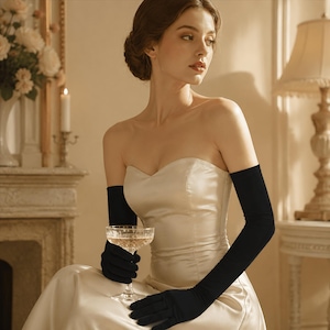 May include: A pair of long, solid black opera-length gloves made of a smooth, opaque fabric. The gloves extend well above the elbow, providing full arm coverage. They are worn with a strapless, cream-colored satin gown with a sweetheart neckline.