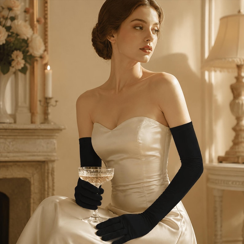 May include: A pair of long, solid black opera-length gloves made of a smooth, opaque fabric. The gloves extend well above the elbow, providing full arm coverage. They are worn with a strapless, cream-colored satin gown with a sweetheart neckline.