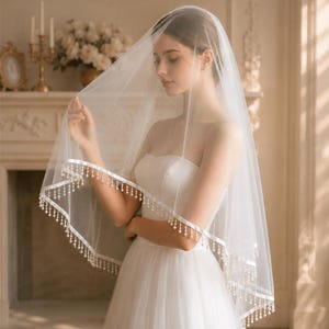 Op de afbeelding: Een doorschijnende witte bruidssluier met een delicate kralenrand. De sluier is gedrapeerd over een witte strapless trouwjurk. De rand heeft kleine, ronde kralen. De algehele uitstraling is elegant en romantisch.