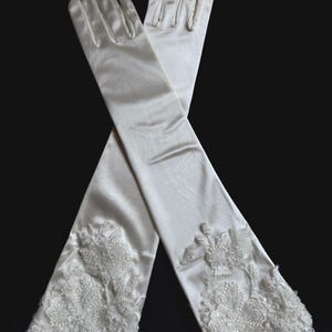 Könnte beinhalten: Ein Paar elfenbeinfarbene, ellenbogenlange Handschuhe. Die Handschuhe bestehen aus einem glatten, satinartigen Material. Die Manschetten sind mit floraler Spitze und Pailletten verziert. Die Handschuhe sind vor einem schwarzen Hintergrund gekreuzt.