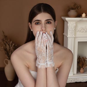 Op de afbeelding: Delicate witte kanten handschoenen met een bloemmotief. De handschoenen hebben een gefranjerde rand bij de pols. De afbeelding toont de ingewikkelde details van het kant en het elegante ontwerp.