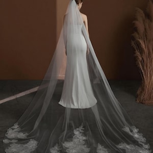 May include: A bride in a white strapless wedding dress, viewed from the back, with a long, flowing veil. The veil features delicate floral lace detailing along the hem. The background is a warm, neutral tone.