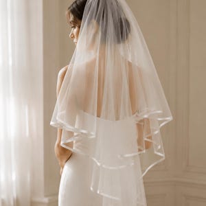 May include: A two-tier white bridal veil featuring a sheer tulle fabric with a smooth satin ribbon trim along the edges. The veil is gathered at the top and drapes down the back, showcasing a layered design with a soft, flowing silhouette.