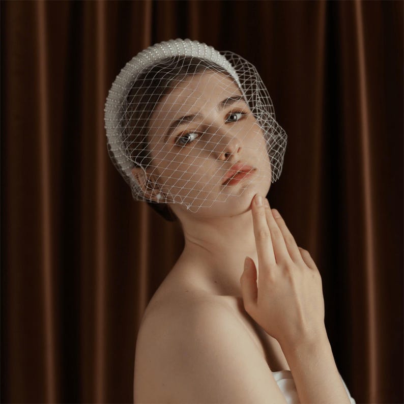May include: A white bridal headband with pearl embellishments and a delicate net veil. The headband is thick and covered in small, round pearls. The veil is a fine, diamond-patterned net that drapes over the face.