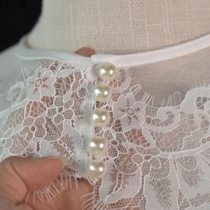 May include: A white garment featuring intricate floral lace detailing across the neckline and upper chest. The piece is fastened with a vertical row of five round, glossy, cream-colored faux pearl buttons, secured by a smooth white fabric trim.