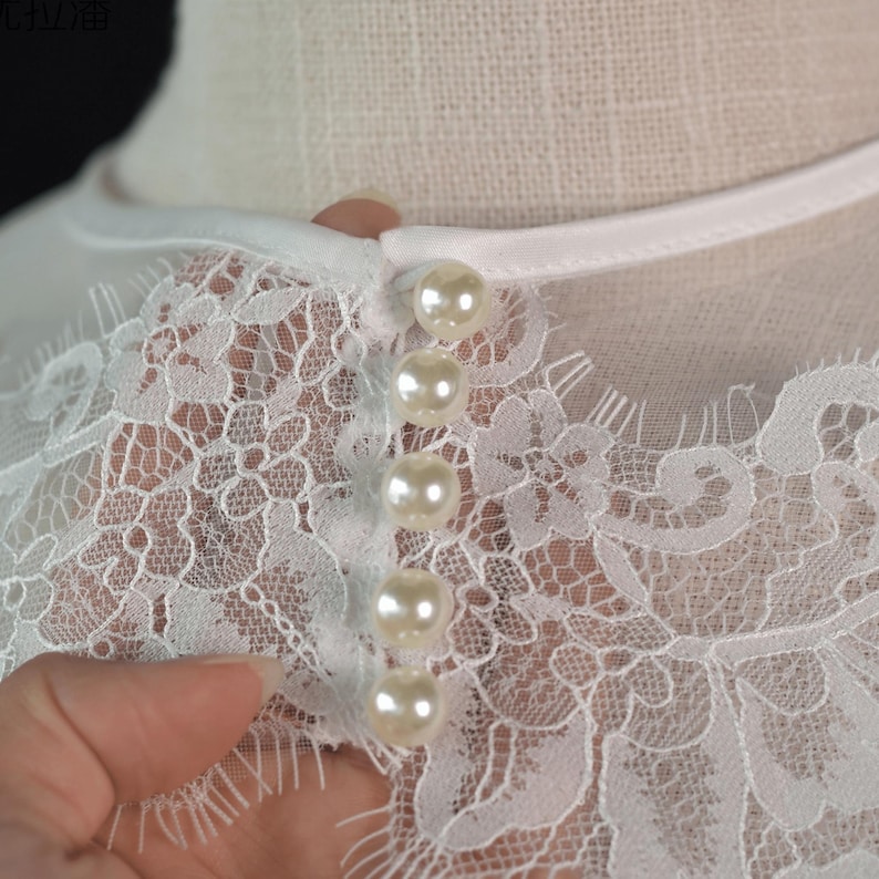 May include: A white garment featuring intricate floral lace detailing across the neckline and upper chest. The piece is fastened with a vertical row of five round, glossy, cream-colored faux pearl buttons, secured by a smooth white fabric trim.