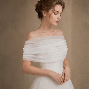 May include: An off-the-shoulder white tulle wrap featuring delicate scattered pearl embellishments. The sheer fabric is layered in soft horizontal folds across the upper chest and shoulders, creating a draped effect over a matching white gown.