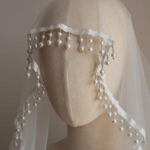 Op de afbeelding: Een doorschijnende witte bruidssluier met een delicate parelrand. De sluier is over een mannequinhoofd gedrapeerd en toont de zachte stof en de elegante pareldetails. De parels zijn langs de rand geregen, wat een vleugje verfijning toevoegt.