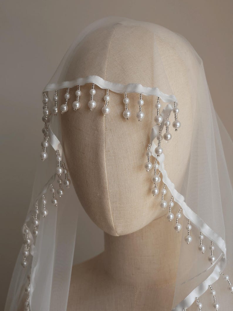 Op de afbeelding: Een doorschijnende witte bruidssluier met een delicate parelrand. De sluier is over een mannequinhoofd gedrapeerd en toont de zachte stof en de elegante pareldetails. De parels zijn langs de rand geregen, wat een vleugje verfijning toevoegt.