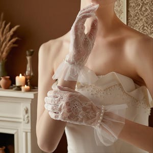 Puede incluir: Delicados guantes de encaje blanco con diseños florales y puños con volantes. Los guantes están adornados con pequeñas perlas en la muñeca, añadiendo un toque de elegancia. La tela transparente permite que se vea la piel, creando un look romántico y refinado.