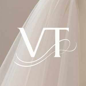以下が含まれることがあります： セリフ体のフォントで「VT」の文字が入った白いロゴ。2本の曲線が添えられています。ロゴは、流れるような白いシアー生地の背景に配置され、柔らかくニュートラルな背景となっています。