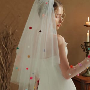 Op de afbeelding: Een witte bruidssluier versierd met veelkleurige pompons. De transparante sluier is versierd met rode, roze, groene en blauwe pompons. De sluier wordt gedragen met een witte strapless jurk. Een bruiloftaccessoire.