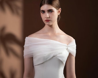 May include: A white off-the-shoulder wedding dress with a draped tulle overlay. The dress features a fitted bodice and a simple, elegant design. The model has her hair pulled back and is wearing small earrings.